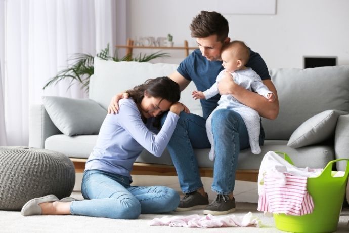 Mulher chorando sentada no chão apoiada no colo de marido que segura bebê 