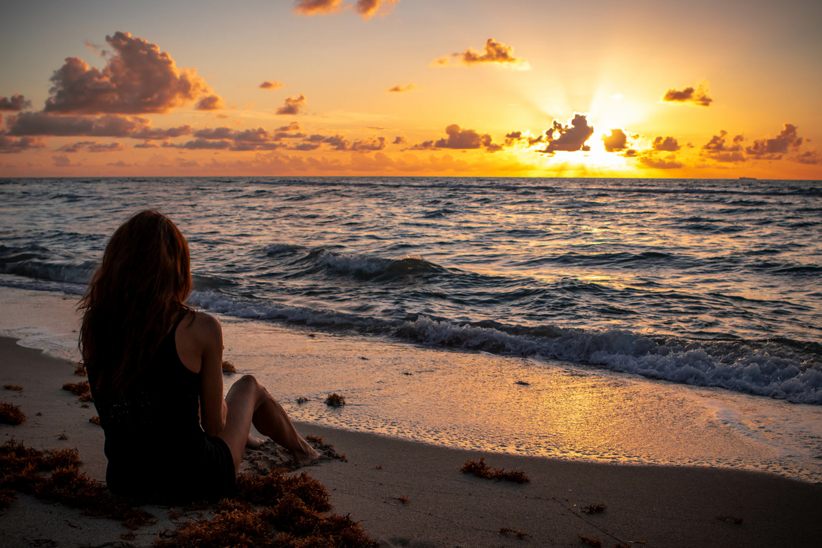 Mulher sentada na beira da praia ao pôr do sol, observando o mar.