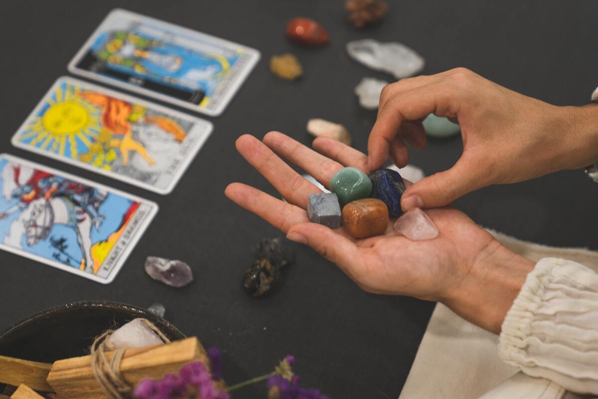 Mão feminina segurando pedras, cartas de Tarot na mesa.