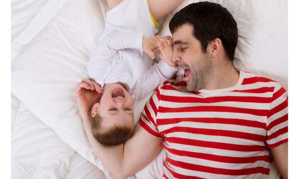 Pai e filho brincando deitados em uma cama.