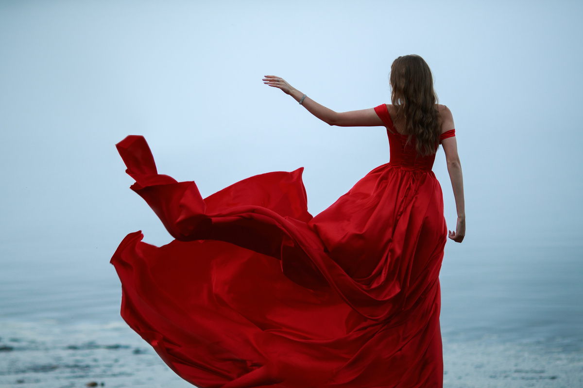 Mulher de costas, jogando saia de vestido vermelho longo ao vento.