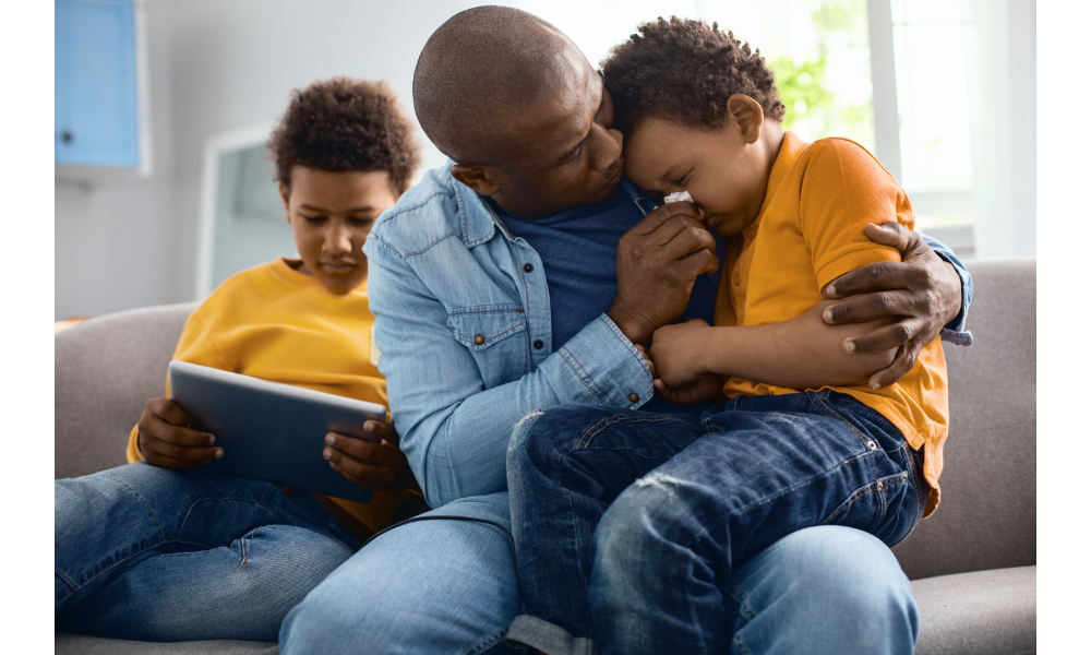 Pai abraçando o filho chorando no colo, ao lado de outro filho em um tablet.