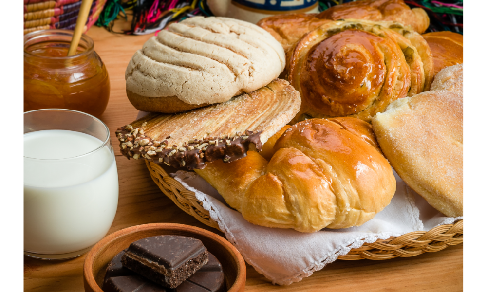 Diversos tipos de pães doces em uma cesta, ao lado de chocolate e um copo com leite.