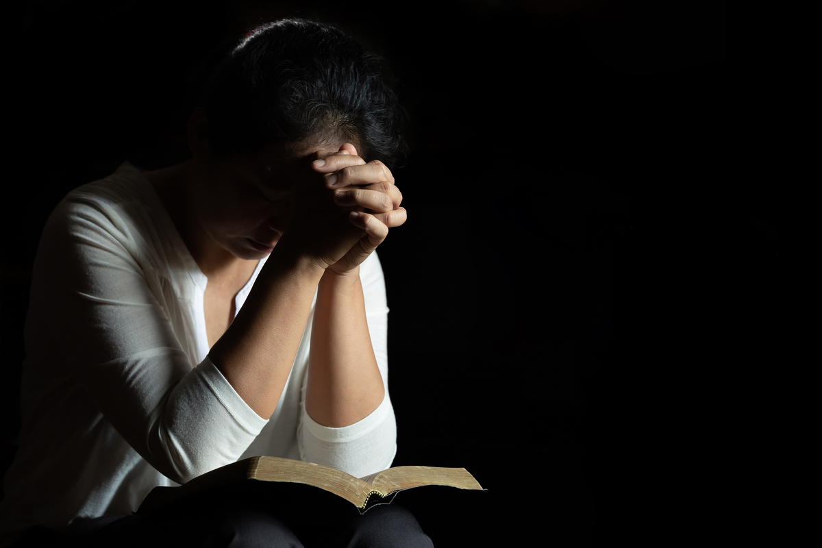 Mulher rezando com Bíblia no colo em ambiente escuro