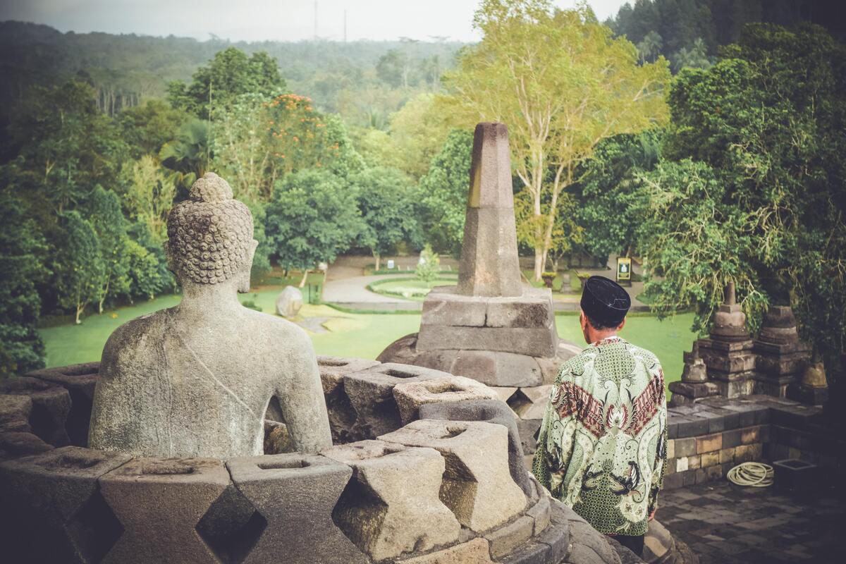 Homem ao lado da figura de Buda, vista de trás. 