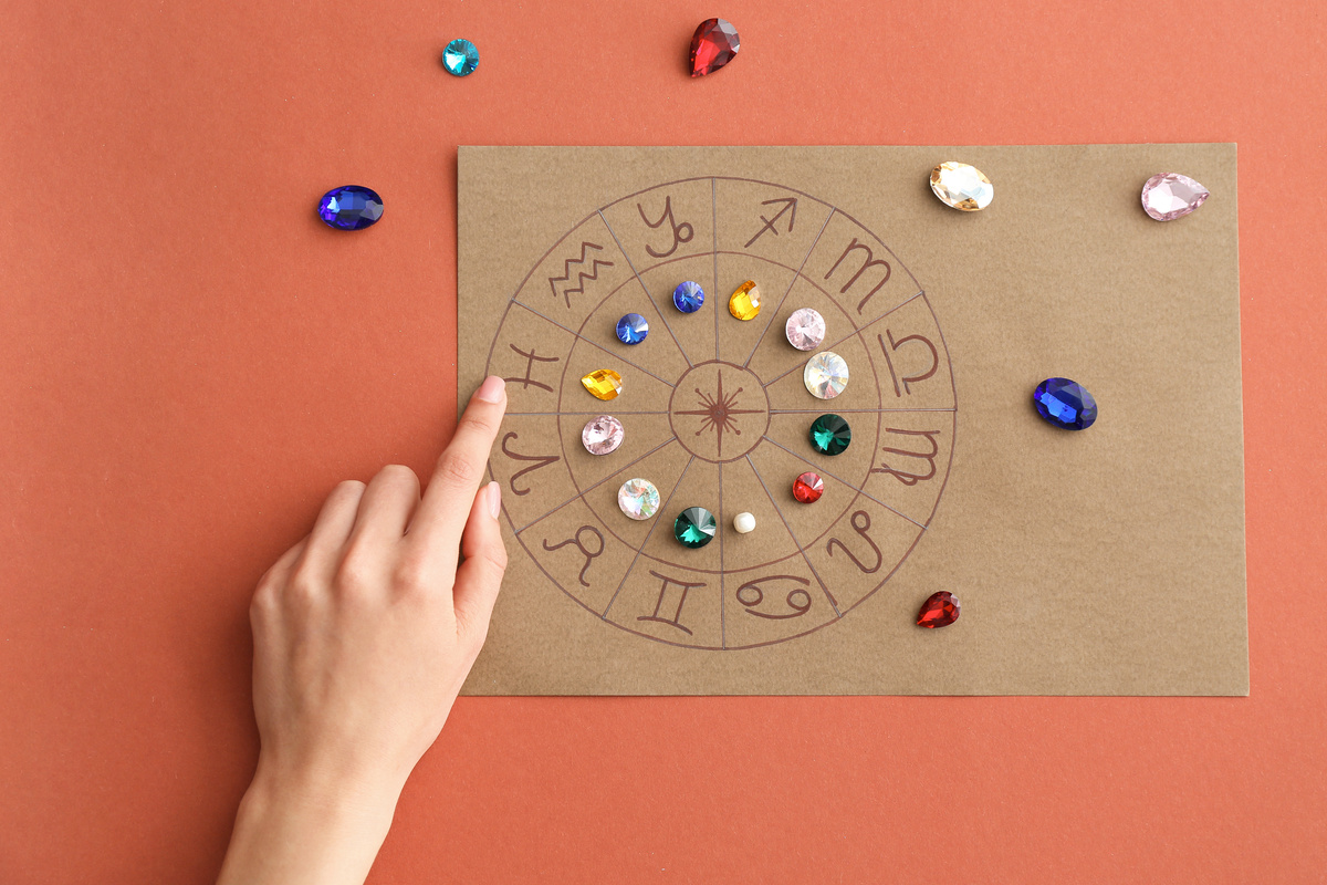 Mão apontando para papel com desenho de círculo do mapa astral, que contém os símbolos dos signos em suas respectivas casas e pedras coloridas em cima, demarcando cada uma delas.