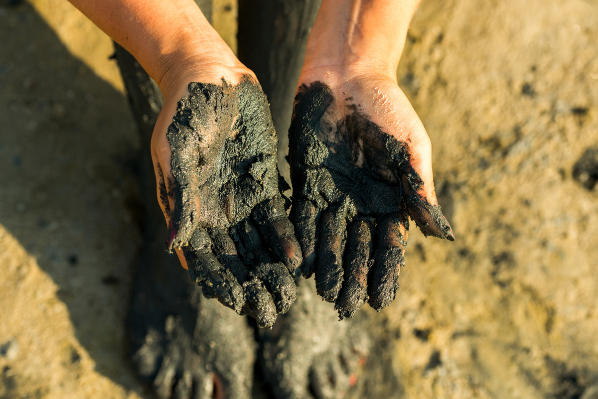 Pessoa mostrando mãos e pés cobertos de lama preta.