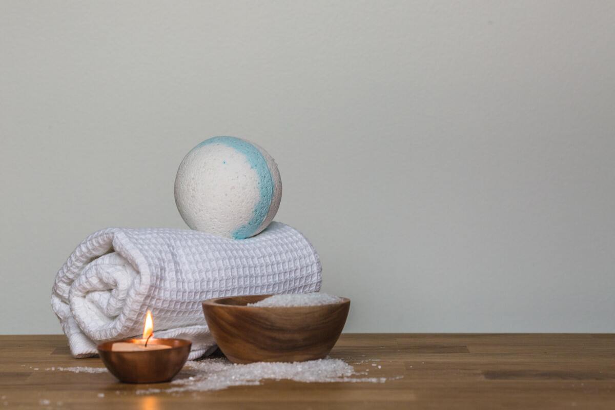 Sal de banho em cima de toalha branca, acompanhado de uma vela acesa e um pote com substância cristalina branca.