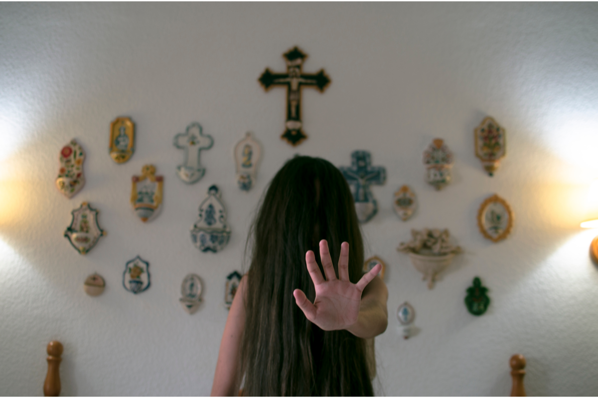 Menina com cabelo na frente do rosto, uma cruz pendurada na parede atrás dela.