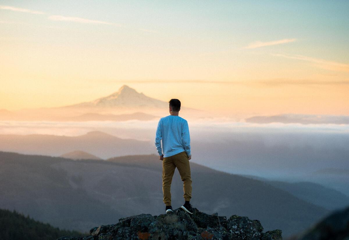 Homem em pé em uma pedra, em frente a uma paisagem de montanhas. 