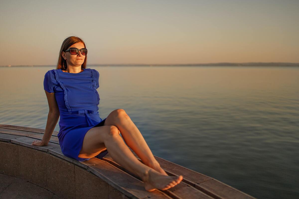 Mulher sentada em uma mureta, em frente ao mar, usando um vestido azul.