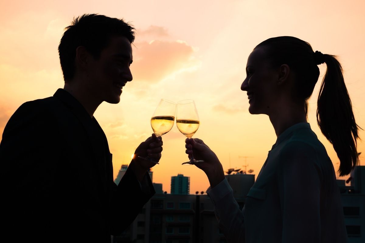 Silhueta do perfil um casal brindando o primeiro encontro com uma taça em um pôr do sol urbano.