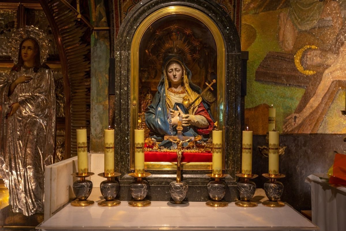 Capela de Nossa Senhora das Dores, que está com as mãos sob o peito. Na foto, há velas douradas acesas na frente da imagem. No centro, Jesus crucificado.