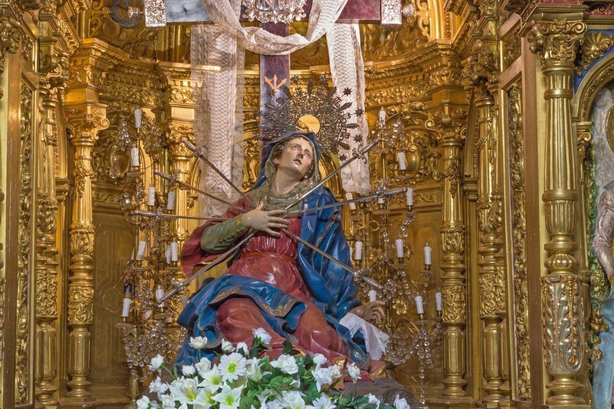 Escultura de Nossa Senhora das Dores em uma igreja com parede dourada e flores nos pés da imagem.