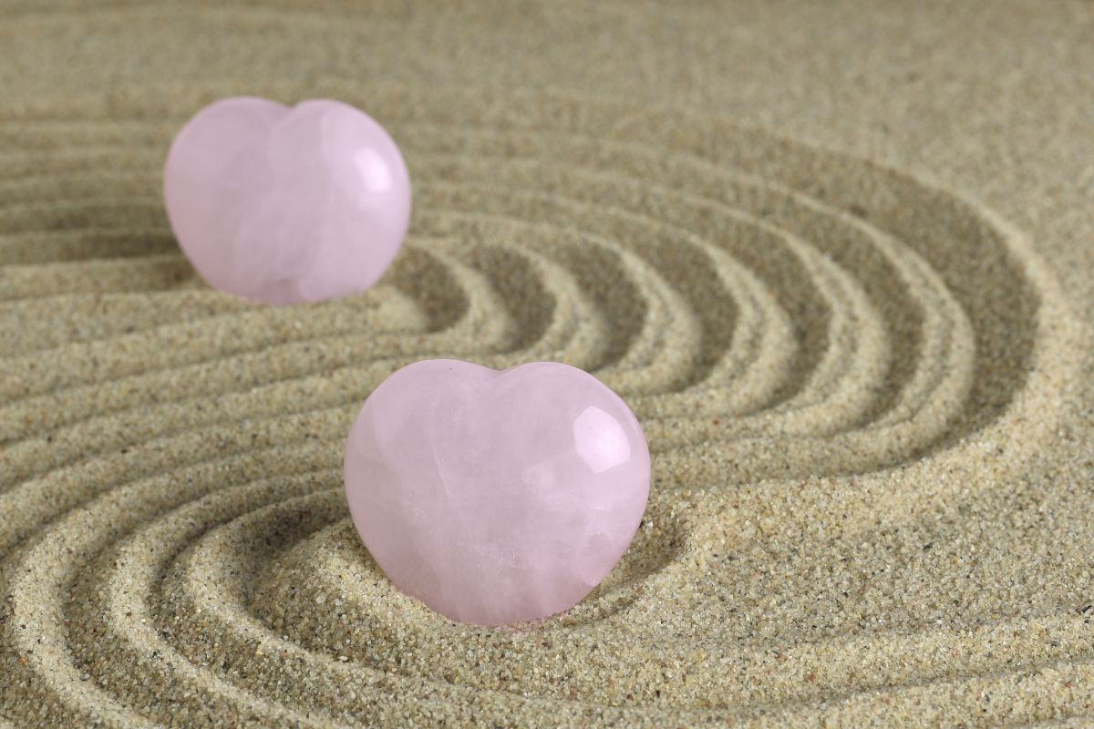 Foto de dois quartzos rosa em formato de coração na areia, desenhando ondas, um atrás do outro.