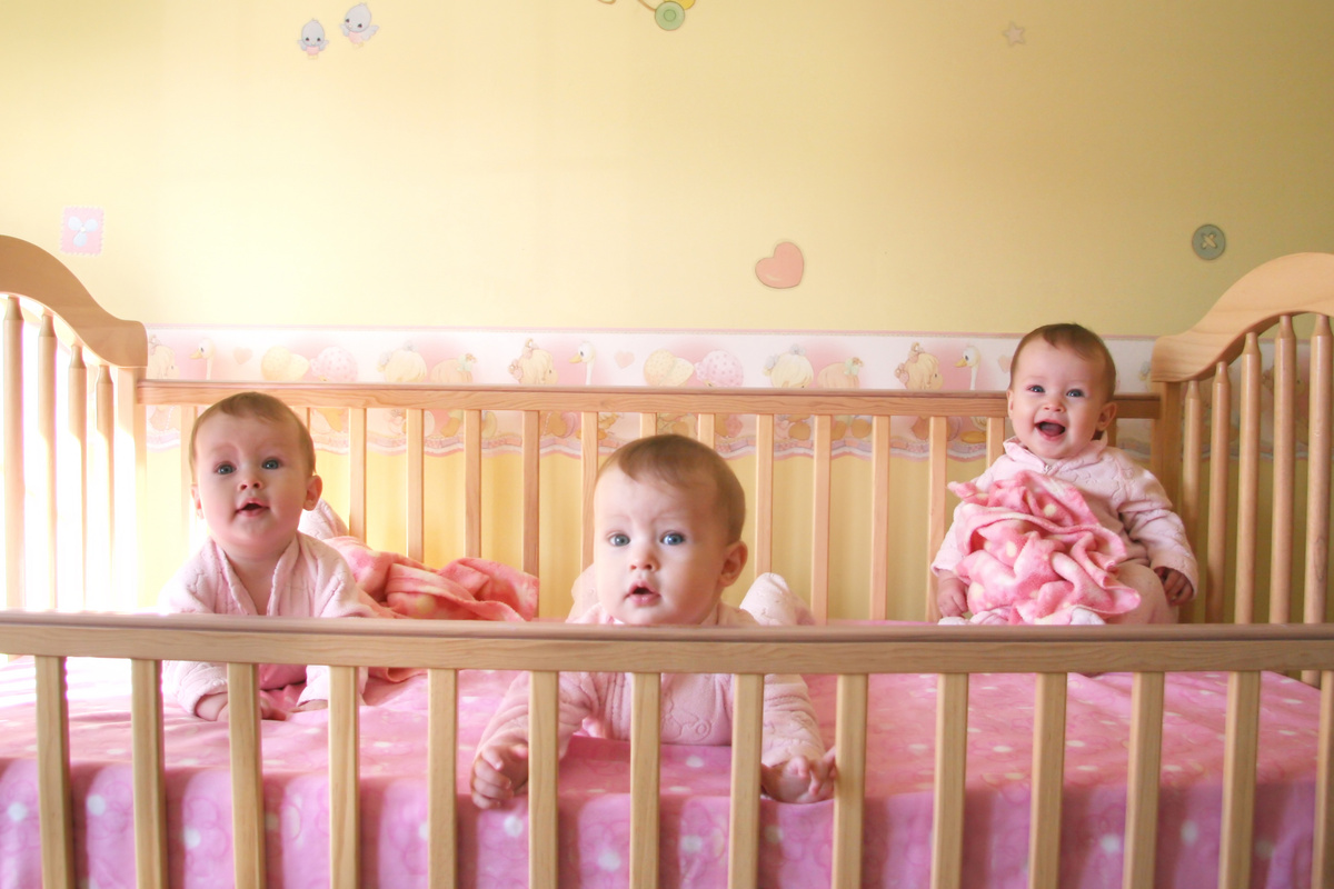 Bebês trigêmeas do sexo feminino deitadas em um único berço, todas vestindo cor-de-rosa enquanto sorriem para cama. 