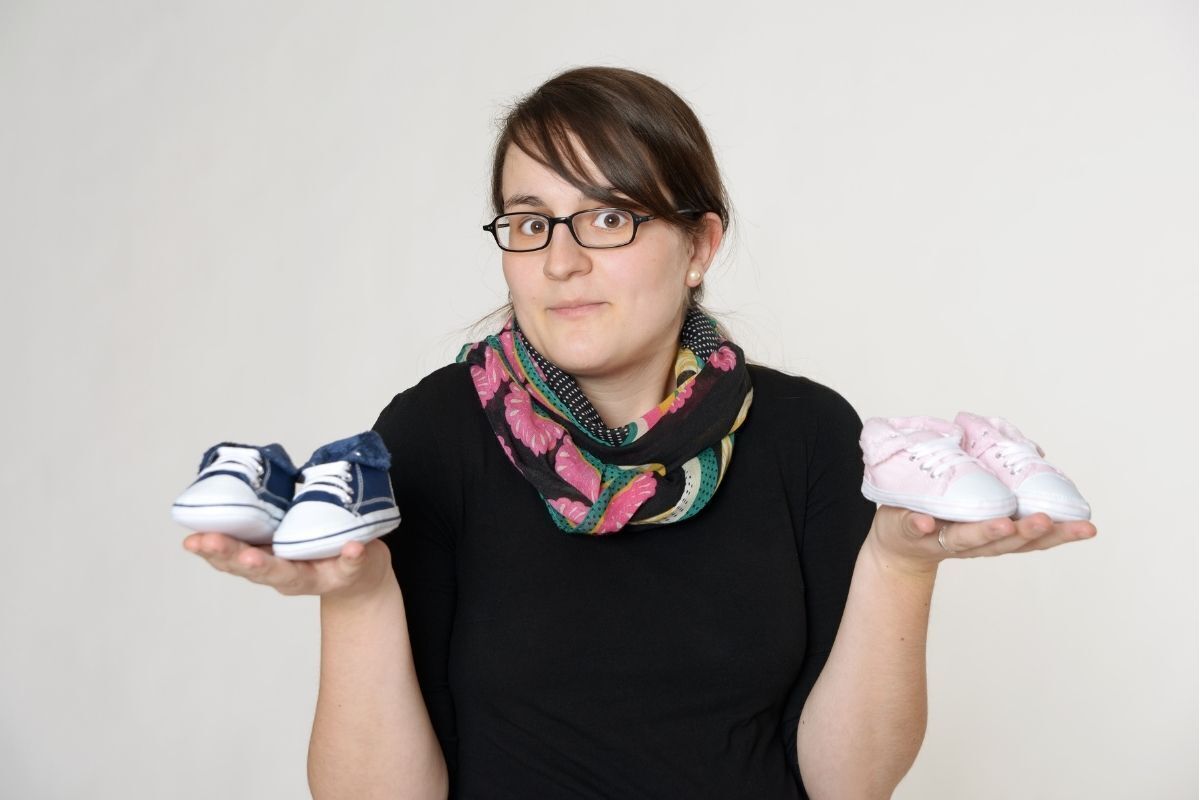 Mulher grávida em dúvida, segurando dois pares de sapatinhos de bebê nas mãos