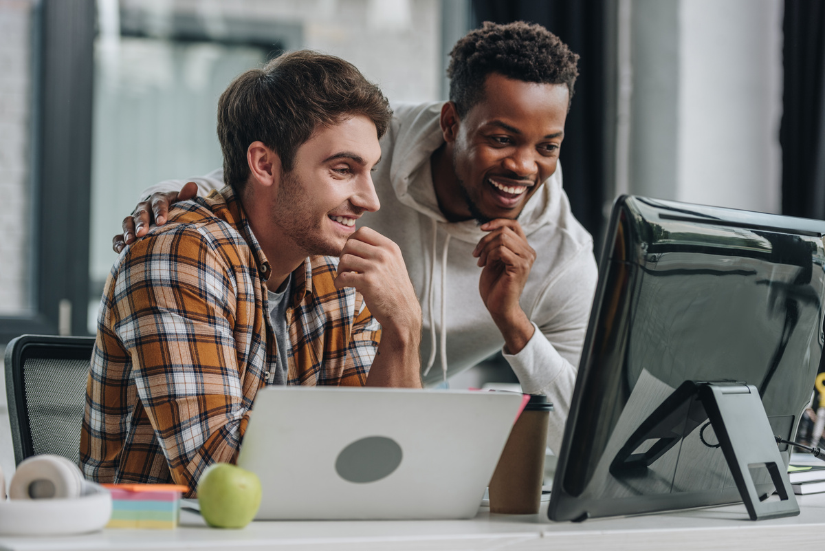 Dois colegas de trabalho sorrindo enquanto visualizam algo juntos em telas de computador, representando a generosidade das pessoas com descendente em Sagitário para com seus colegas de trabalho.