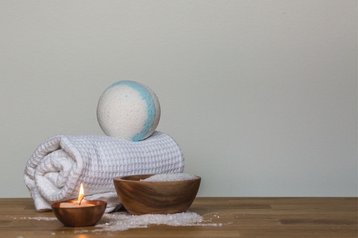 Imagem aconchegante de toalha branca enrolada em uma mesa de madeira, com uma vela branca acesa e sal grosso em um recipiente.