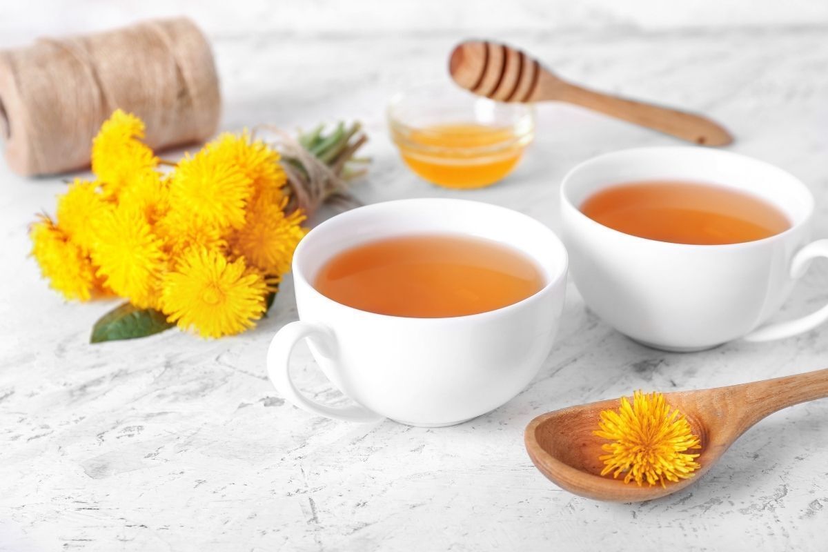 Chá de dente-de-leão, com flores amarelas em uma xícara branca e uma espátula de madeira ao lado