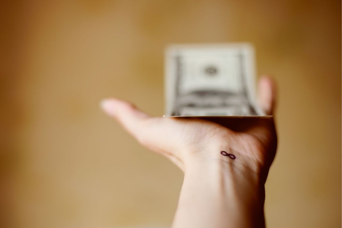 Uma pessoa está segurando notas de dinheiro de papel em sua mão, com uma tatuagem do símbolo do infinito em seu pulso