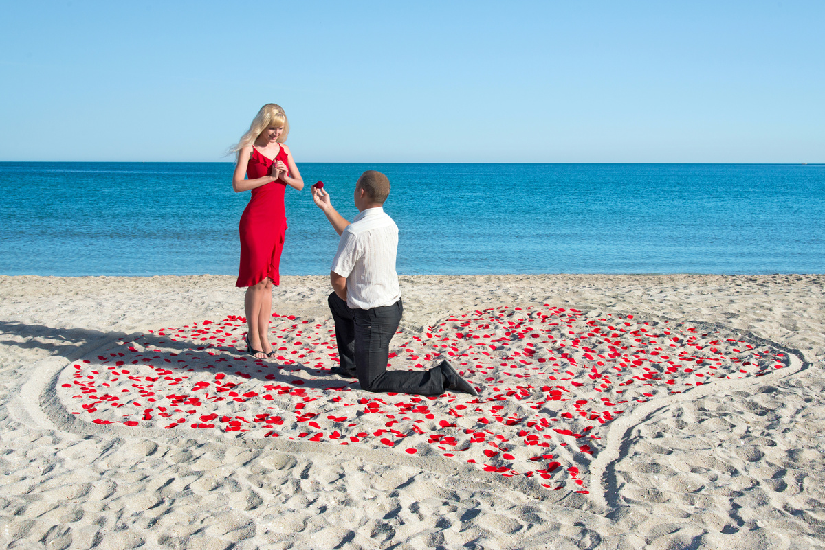 Homem se declarando para mulher na praia, ajoelhado em um coração de pétalas de rosas