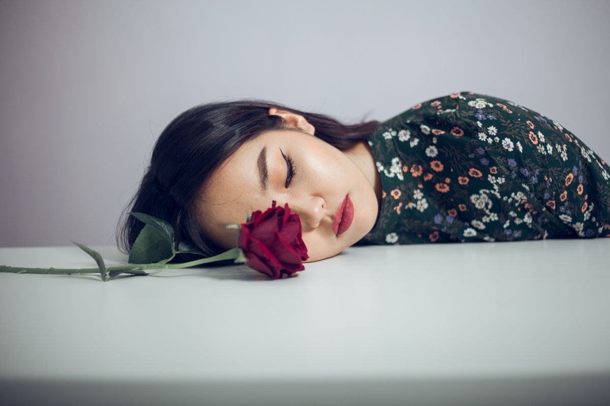 Fotografia ilustrativa de uma mulher deitada sob a mesa com uma rosa vermelha ao seu lado. Ela está com os olhos fechados. 