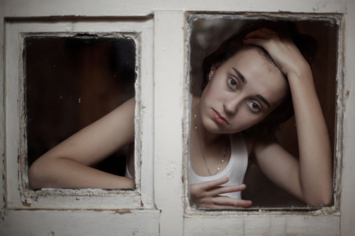 Menina na parte interna de uma casa com uma mão na cabeça e olhar melancólico, sendo vista pela janela.