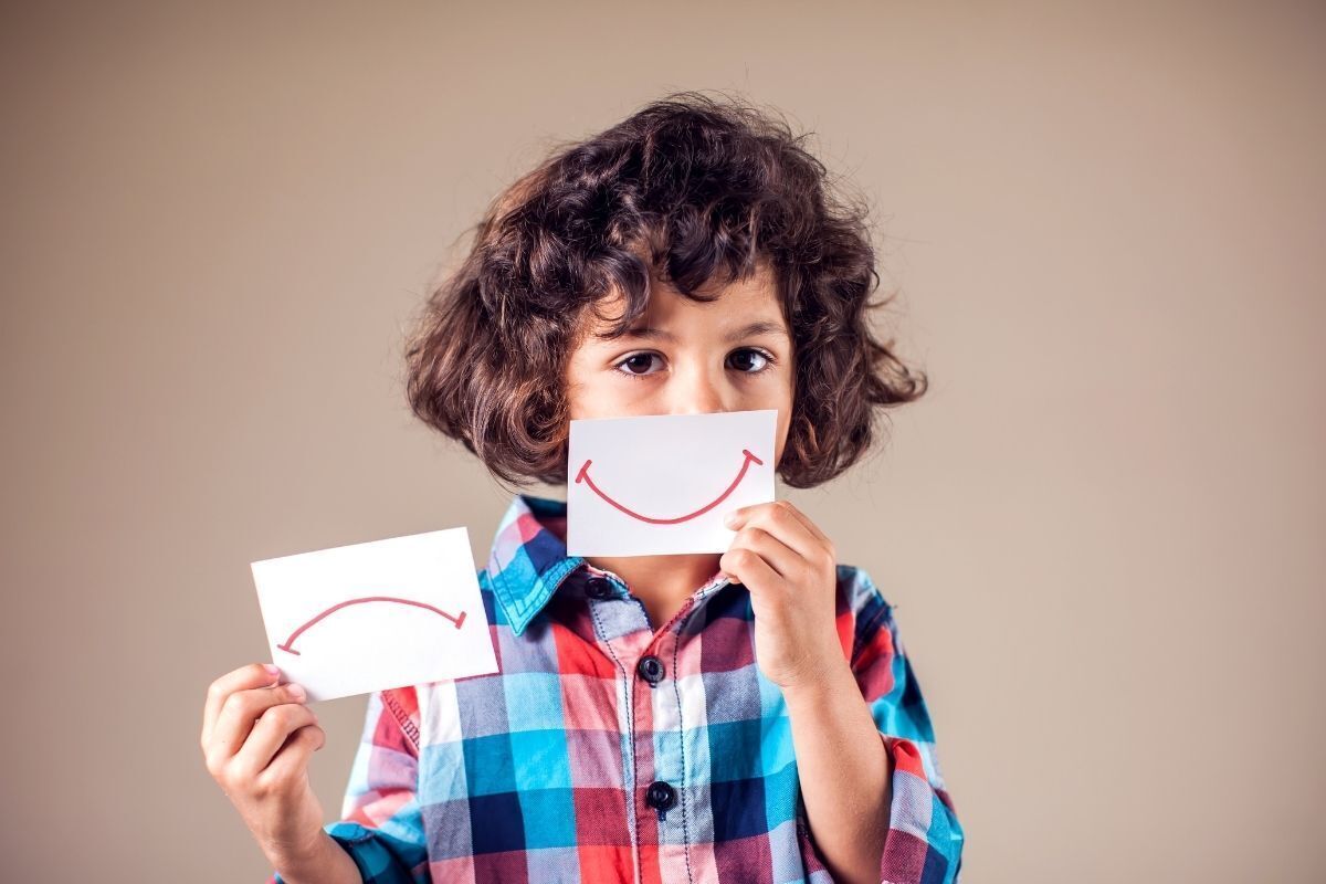 Fotografia de uma criança segurando um cartão com o desenho de um sorriso na frente da sua boca. Em outra mão, segura um cartão com uma boca desenhada, representando a tristeza.