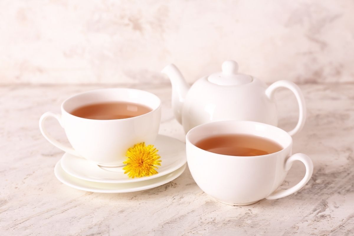 Duas xícaras de chá com pires branco. Uma delas tem a flor dente-de-leão da cor amarela para decoração.