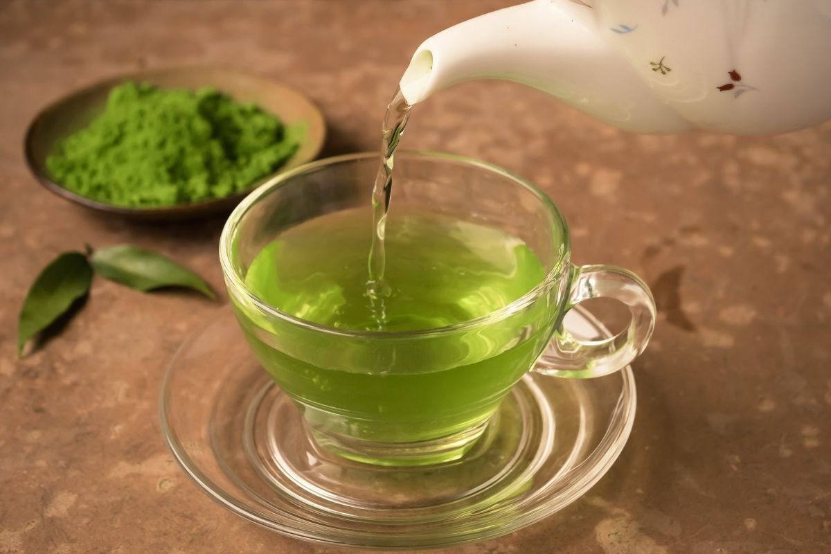 Chá verde sendo servido em uma xícara transparente. Ao fundo, o pó verde desse chá aparece em um recipiente. 
