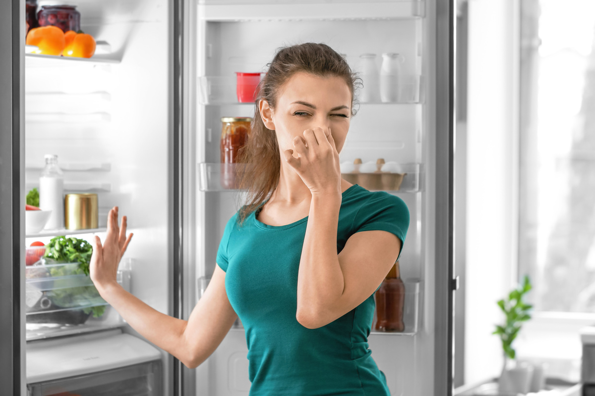 Mulher tampa o nariz e vira o rosto ao abrir a geladeira e se deparar com comida estragada