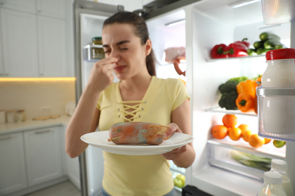 Mulher retirando comida estragada da geladeira e tampando o nariz para não sentir o cheiro