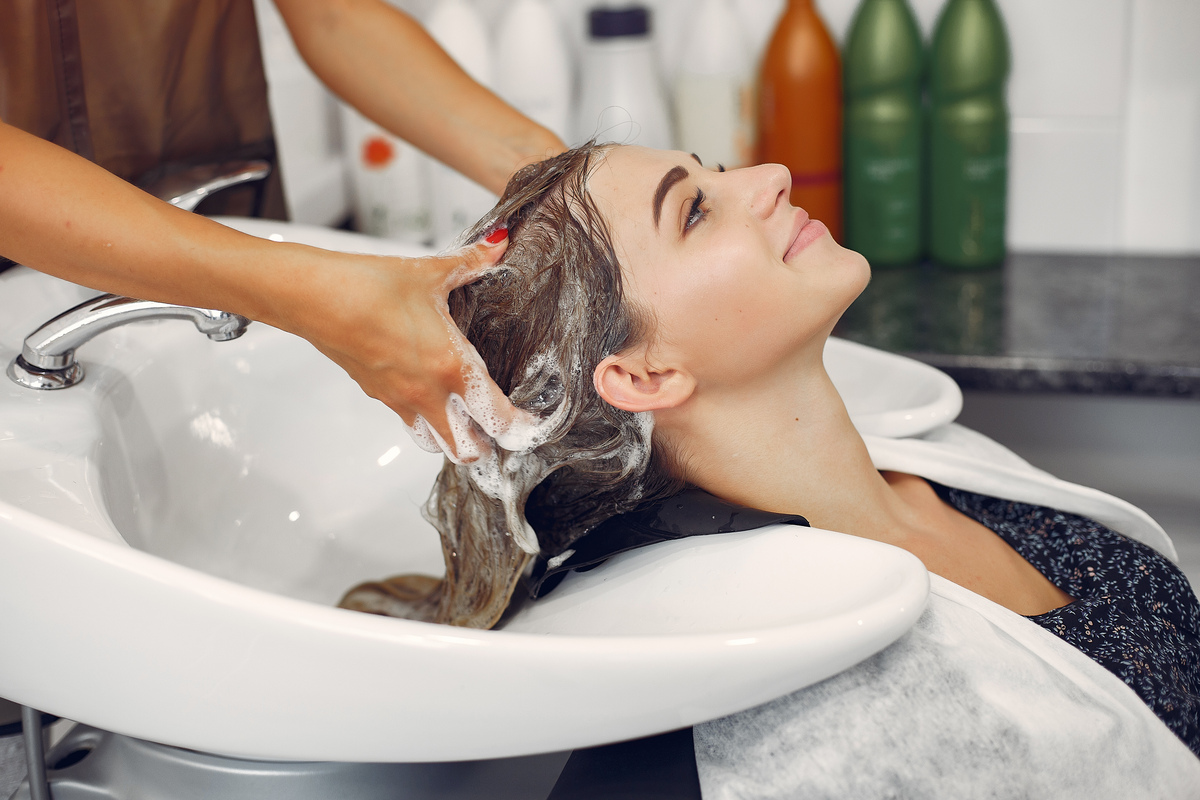Mulher sentada em tanque de cabeleireiro, enquanto tem seu cabelo lavado por outra pessoa.