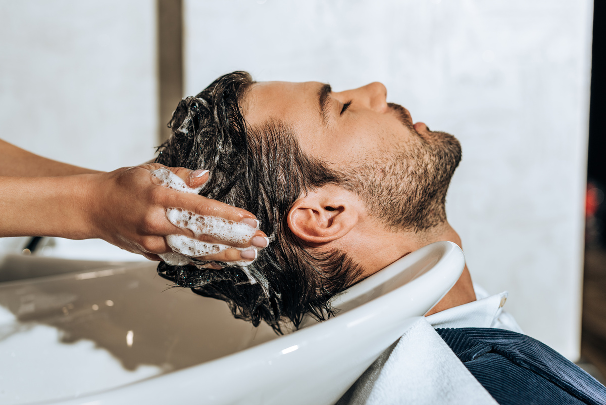 Homem sentado com a cabeça em tanque, enquanto outra pessoa lava e massageia o seu cabelo.
