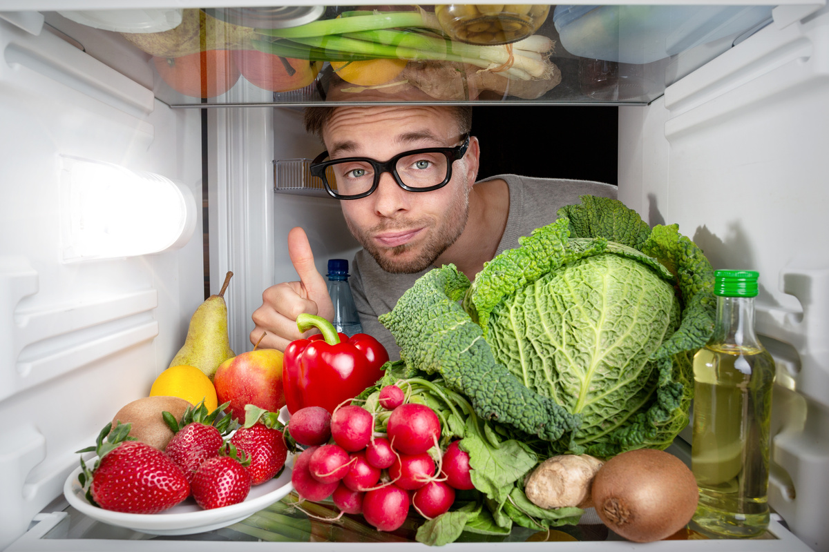 Homem fazendo sinal de sim com a mão enquanto olha para geladeira cheia de alimentos naturais