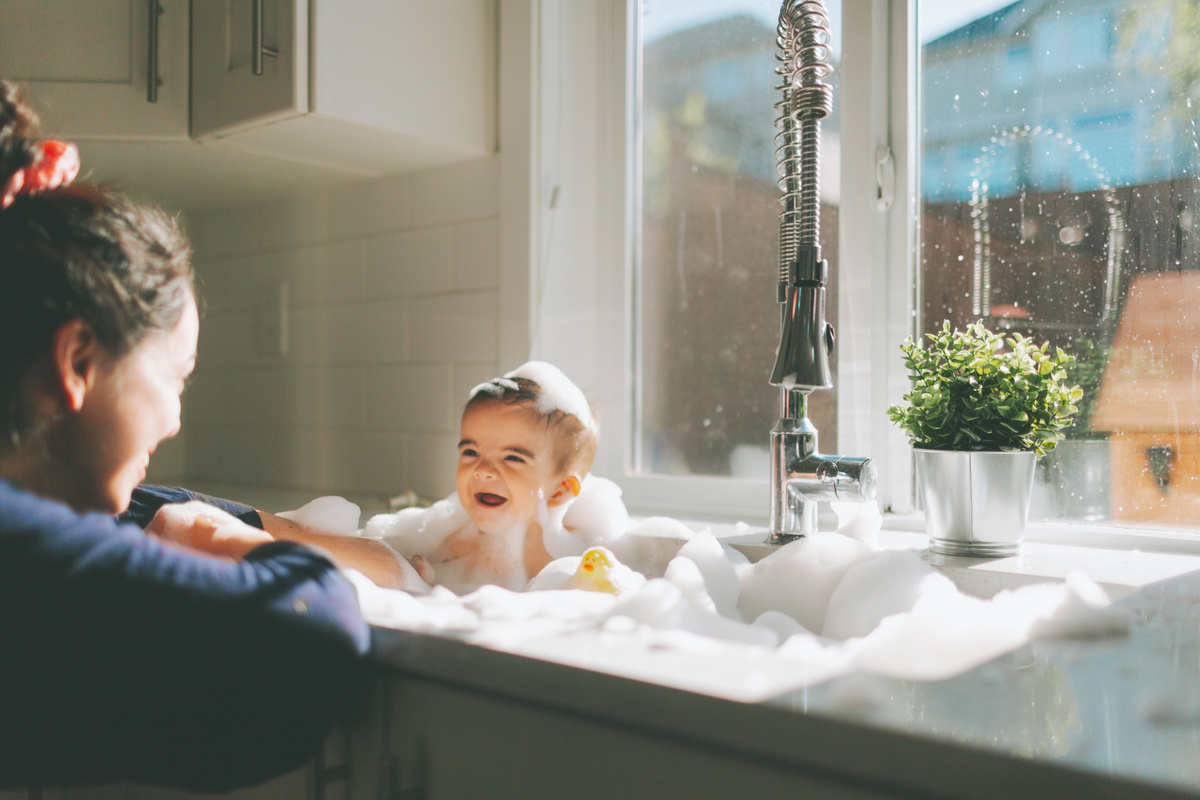 Mulher dando banho em bebê, que sorri para ela enquanto se senta em banheira cheia de espuma.