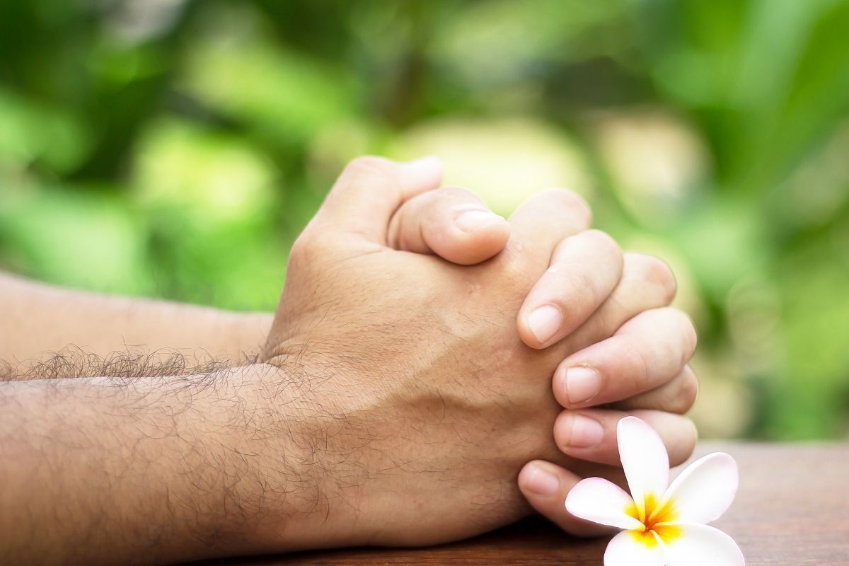 Mãos entrelaçadas em forma de oração e uma flor branca ao lado