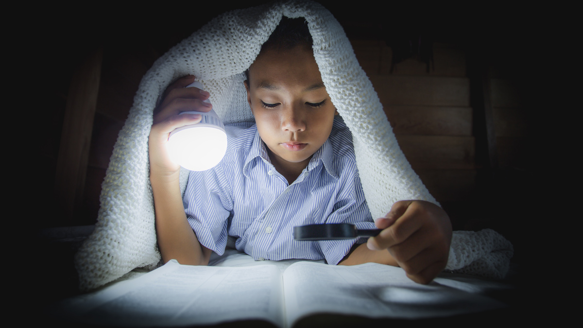 Criança lendo Bíblia com lanterna e lupa embaixo das cobertas