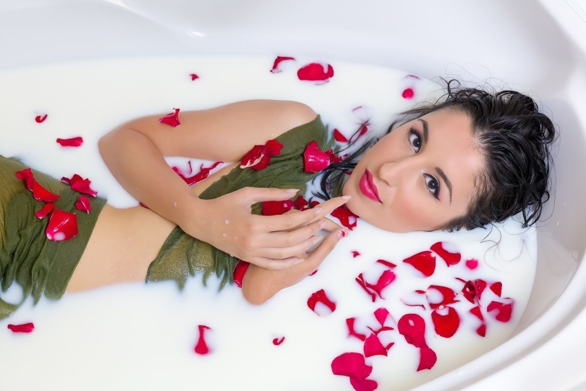 Mulher vestida de verde deitada em uma banheira com leite e pétalas de rosas