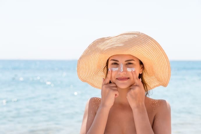 Mulher na praia com chapéu passando protetor solar no rosto