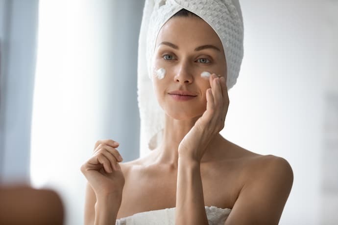 Mulher com toalha prendendo os cabelos passando ácido hialurônico no rosto