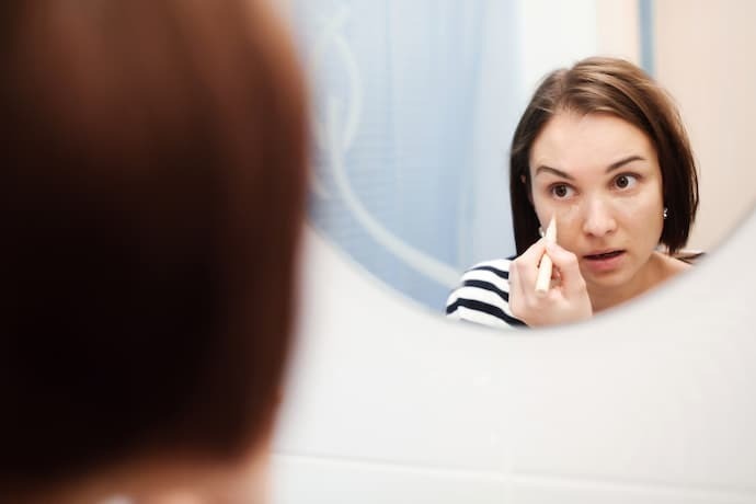 Reflexo de mulher no espelho passando corretivos para olheiras