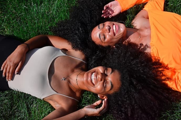 Duas mulheres com cabelos cacheados rindo deitadas na grama