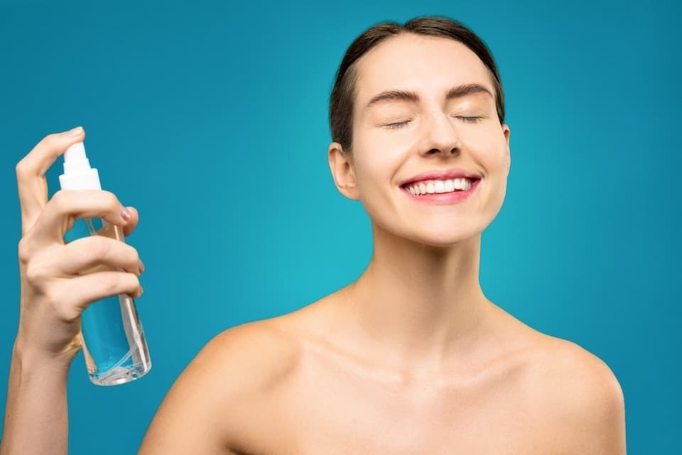 Mulher sorridente em fundo azul aplicando água micelar no rosto