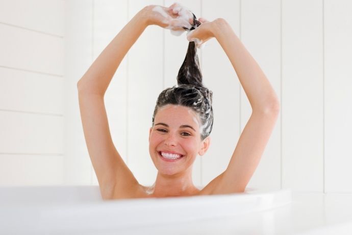 Mulher sorridente na banheira lavando os cabelos com shampoo