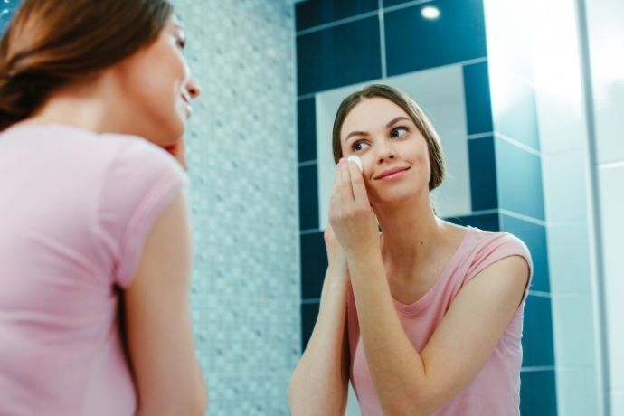 Mulher se olhando no espelho enquanto passada algodão no rosto com adstringente