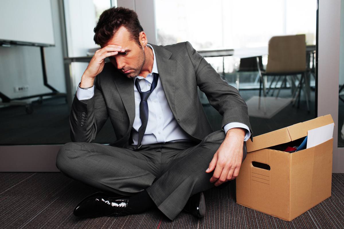 Homem sentado no chão, junto de seus materiais de escritório, lamentando a própria demissão.