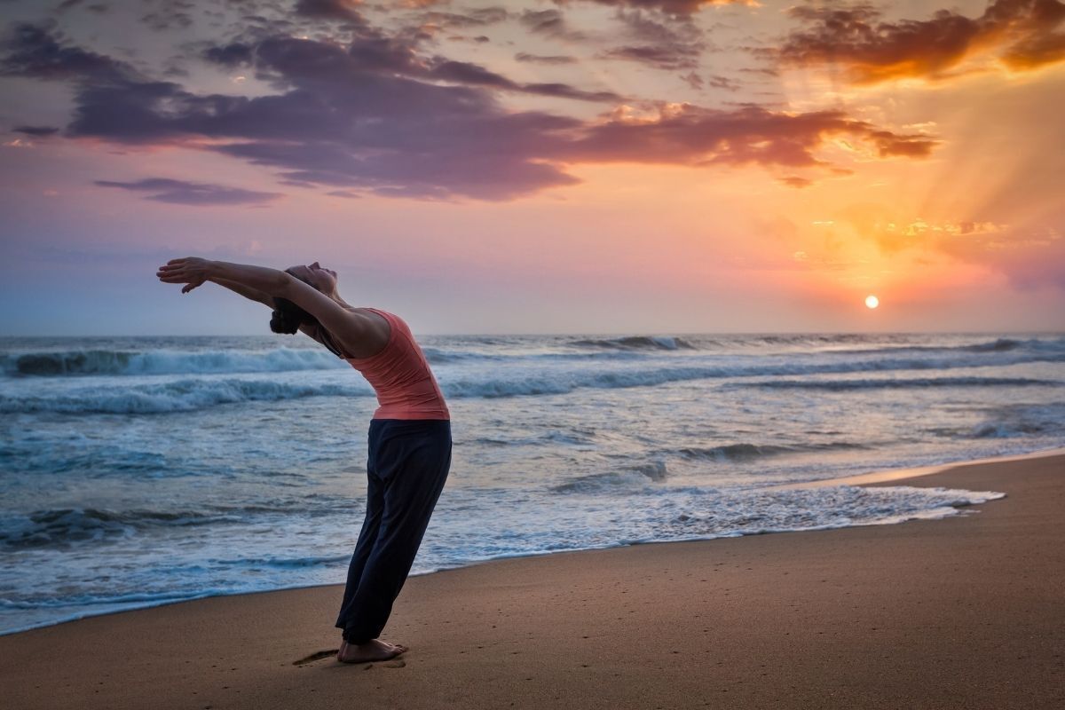 Mulher praticando movimentos do Surya Namaskar na praia, durante o nascer do sol.