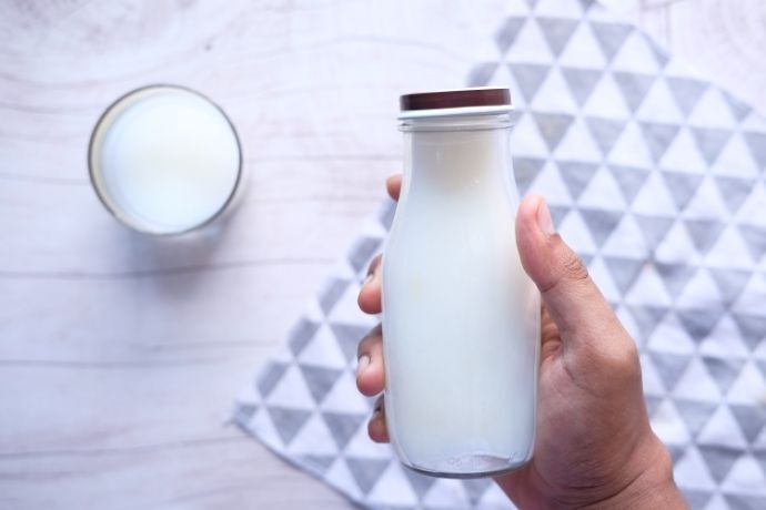 Pessoa segurando garrafa de vidro com leite e copo de leite ao fundo